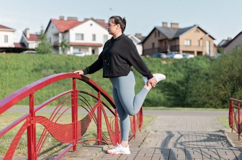 A woman in active wear performs a stretching exercise on a red bridge in a suburban park setting.