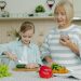 Grandmother with smartphone is teaching granddaughter to cook salad at home, cute little girl is cutting cucumber using knife and chopping board.
